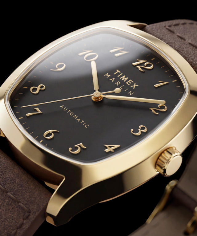 Close-up van de goudkleurige Timex Marlin Chatham Automatic met zijn zwarte wijzerplaat, opgelegde goudkleurige Arabische cijfers, goudkleurige wijzers en bruine leren band
