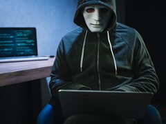 Een gemaskerde figuur met capuchon werkt op een laptop in een verduisterde kamer.