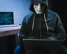 Een gemaskerde figuur met capuchon werkt op een laptop in een verduisterde kamer.