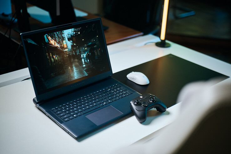 Een open Alienware-gaminglaptop, opgesteld op een wit studiotafelblad, waarop een donkere, reflecterende straat scène wordt weergegeven met scherpe, path-traced details, gecombineerd met een zwarte controller en een witte draadloze muis.