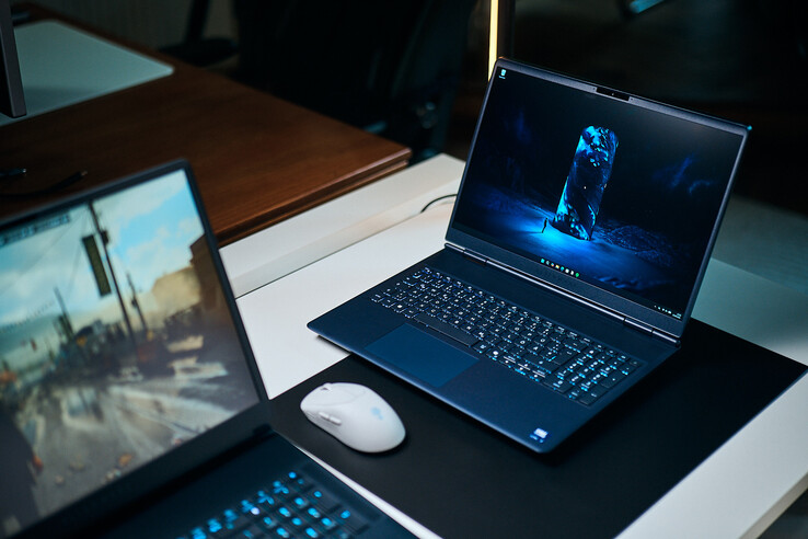 Een donkerblauwe gaming-laptop uit het middensegment (Alienware Aurora 16X) staat schuin op een bureauonderlegger met een gloeiend blauwe scifi-achtergrond, naast een witte draadloze muis, met op de voorgrond een andere laptop die wazig is weergegeven.