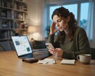 Een vrouw zit aan een bureau en kijkt naar haar smartphone; ernaast liggen een laptop, bonnetjes en een notitieblok