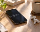 Smartphone die een audiogolfvorm weergeeft naast draadloze oordopjes en een koffiekopje op een houten bureau