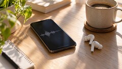 Smartphone die een audiogolfvorm weergeeft naast draadloze oordopjes en een koffiekopje op een houten bureau