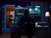 Een onderzoeker met capuchon werkt op meerdere schermen in een verduisterde opstelling. Generieke hacker afgebeeld. 