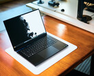 Een open, matzwarte Razer Blade 16-gaminglaptop die op een witte bureauonderlegger op een warme houten tafel staat, met een levendige zwart-witte achtergrondafbeelding.