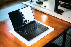 Een open, matzwarte Razer Blade 16-gaminglaptop die op een witte bureauonderlegger op een warme houten tafel staat, met een levendige zwart-witte achtergrondafbeelding.