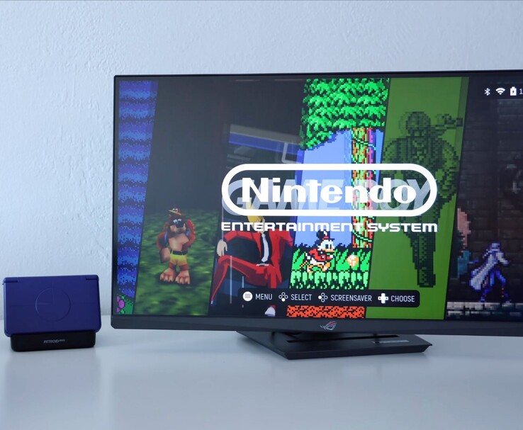 Retroid Pocket Flip 2 in paars geplaatst op een dock naast een monitor, met op het TV-scherm een retro NES-emulatorinterface. (Afbeelding bron: Retro Game Corps op YouTube)