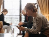 Een tiener kijkt naar haar smartphone, terwijl op de achtergrond twee volwassenen onscherp zitten.
