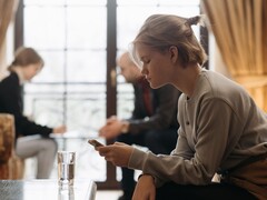 Een tiener kijkt naar haar smartphone, terwijl op de achtergrond twee volwassenen onscherp zitten.