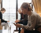 Een tiener kijkt naar haar smartphone, terwijl op de achtergrond twee volwassenen onscherp zitten.