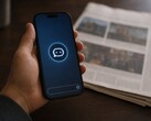 Een hand houdt een smartphone vast waarop een chatbot-pictogram te zien is; ernaast ligt een gedrukte krant.