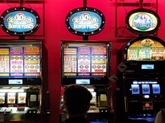 Speelautomaten in een casino met helder verlichte displays en spelende mensen ervoor (Afbeelding bron: djedj via Pixabay)