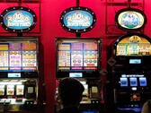 Speelautomaten in een casino met helder verlichte displays en spelende mensen ervoor (Afbeelding bron: djedj via Pixabay)
