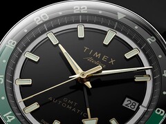 Close-up van de zwarte wijzerplaat van de Timex Atelier GMT Première M1a, met goudkleurige wijzers, lichtgevende uurmarkeringen, een datumvenster en een zwart-groene 24-uurs-GMT-ring.