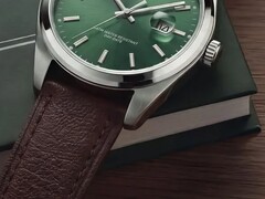 Close-up van het Timex Legacy Day and Date-horloge, met zijn groene wijzerplaat met sunray-afwerking, zilverkleurige lunette en geborstelde bandaanzetten, geribbelde kroon en bruine leren band met textuur, tegen de achtergrond van een donkergroene doos en een houten oppervlak.
