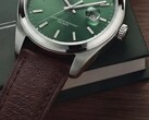 Close-up van het Timex Legacy Day and Date-horloge, met zijn groene wijzerplaat met sunray-afwerking, zilverkleurige lunette en geborstelde bandaanzetten, geribbelde kroon en bruine leren band met textuur, tegen de achtergrond van een donkergroene doos en een houten oppervlak.