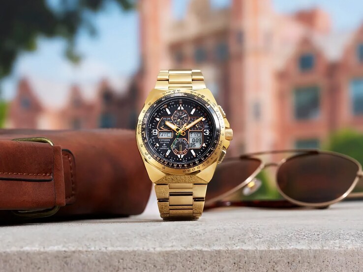 Een goudkleurig Citizen Promaster Skyhawk A-T JY8144-50E pilotenhorloge staat rechtop op een richel naast een bruine leren tas en een zonnebril, met op de achtergrond een wazig stadsgezicht.