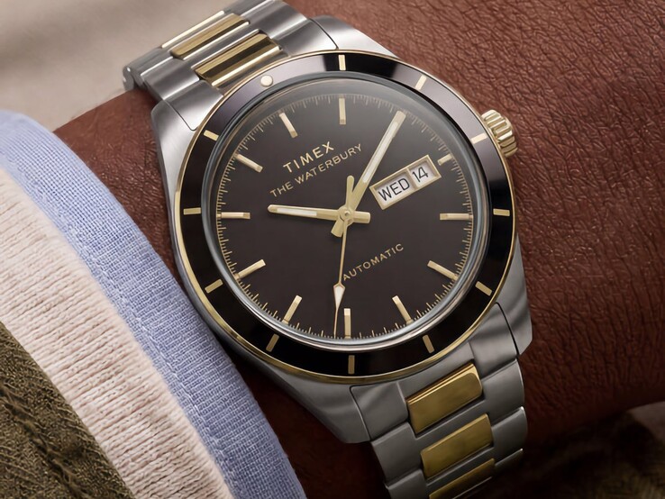 Een tweekleurig Timex Waterbury Heritage Automatic-horloge met zwarte wijzerplaat, gedragen om de pols, waarop de dag- en datumweergave en de goudkleurige accenten te zien zijn.