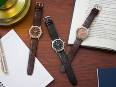 Drie Citizen Rolan-horloges, neergelegd op een houten bureau naast een notitieboekje, een pen, een lamp en een open boek, waaronder het model AW2110-14Z met zalmkleurige wijzerplaat.