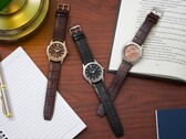 Drie Citizen Rolan-horloges, neergelegd op een houten bureau naast een notitieboekje, een pen, een lamp en een open boek, waaronder het model AW2110-14Z met zalmkleurige wijzerplaat.