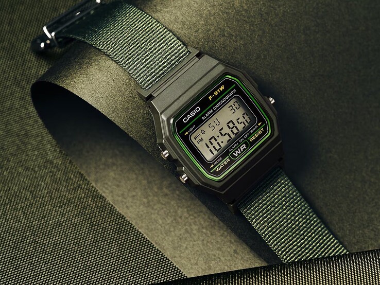 Casio F-91WF-3A digitaal horloge met een zwarte kast met groene accenten en een geweven groene stoffen band, gefotografeerd tegen een donkergroene achtergrond met textuur.