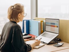 Een vrouw met een bril werkt aan een spreadsheet op een laptop, met ordners en een rekenmachine op het bureau