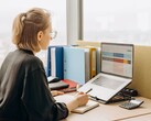 Een vrouw met een bril werkt aan een spreadsheet op een laptop, met ordners en een rekenmachine op het bureau