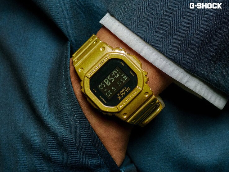 Close-up van een gouden Casio G-Shock DW-5600BEP-9-horloge dat om de pols wordt gedragen tegen een donkerblauwe achtergrond. Het horloge heeft een zwart digitaal display, een gouden kast en band, en het BlackEyePatch-logo op de lunette en de wijzerplaat.