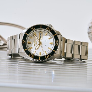 Seiko 5 Sports SKX HDB004 horloge met een zilverwitte wijzerplaat, zwarte draaibare lunette met goudkleurige markeringen, goudkleurige wijzers en indexen, en een roestvrijstalen horlogeband, schuin gefotografeerd.