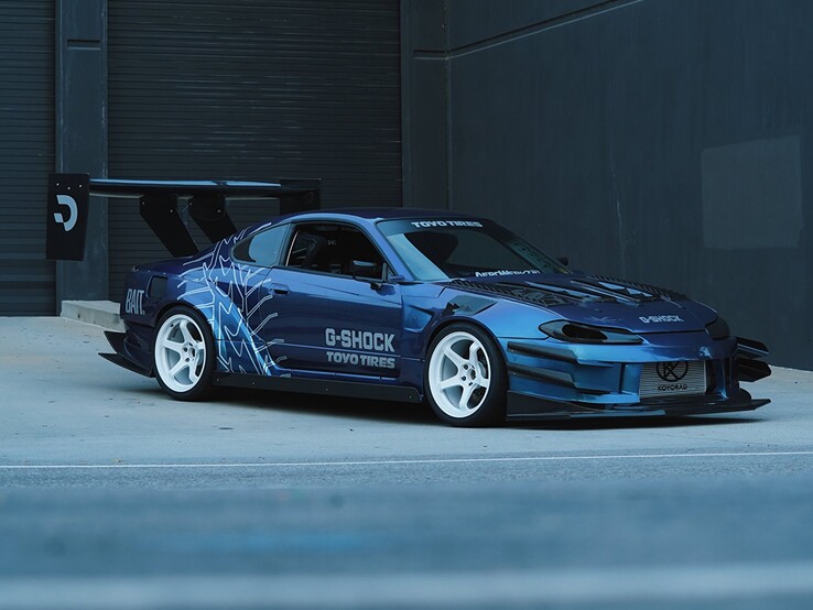 Blauwe driftauto met afbeeldingen van G-Shock en Toyo Tires geparkeerd voor een industrieel gebouw.