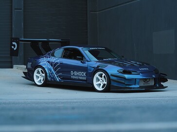 Een blauwe driftwagen met afbeeldingen van G-Shock en Toyo Tires, geparkeerd voor een bedrijfsgebouw.