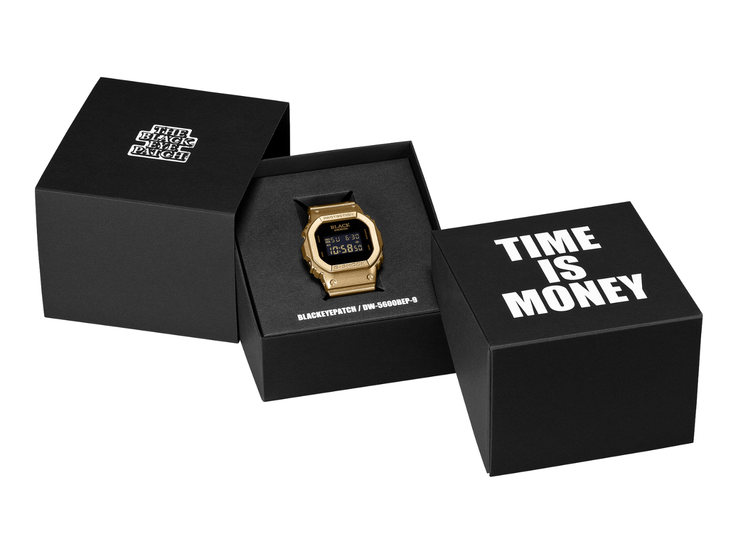 Een gouden Casio G-Shock DW-5600BEP-9-horloge met een zwart digitaal display ligt tussen twee zwarte presentatiedozen in. Op de wijzerplaat van het horloge staat het BlackEyePatch-logo, terwijl op de doos op de voorgrond „TIME IS MONEY“ staat.
