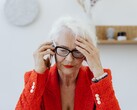 Een oudere vrouw met wit haar en een bril, die in een lichte woonkamer aan de telefoon is en haar hand tegen haar slaap houdt.