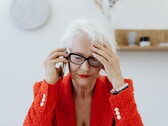 Een oudere vrouw met wit haar en een bril, die in een lichte woonkamer aan de telefoon is en haar hand tegen haar slaap houdt.