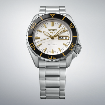 Seiko 5 Sports SKX HDB004 met een zilverwitte wijzerplaat, zwarte bezel met goudkleurige markeringen, goudkleurige wijzers en indexen, en een roestvrijstalen band, afgebeeld tegen een lichtgrijze achtergrond.